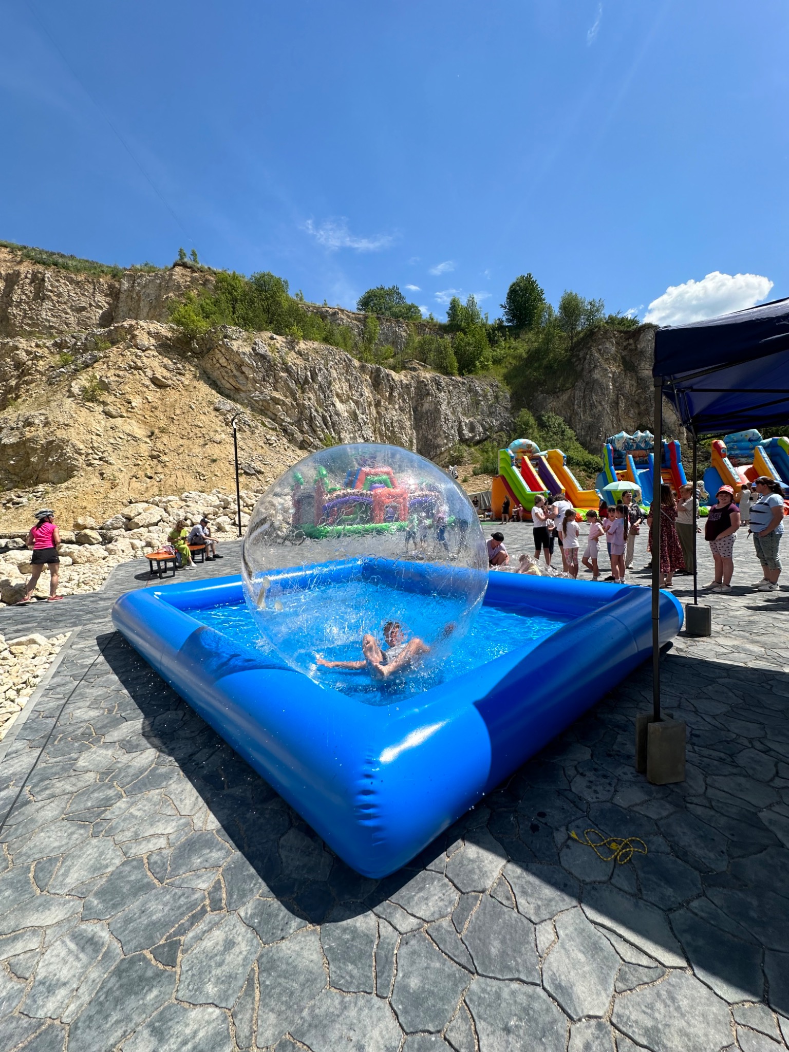 Atrakcja wodna w Michałowicach: osoba w kuli wodnej na tle dmuchańców i skał. Błękitny basen, słońce i bawiący się ludzie tworzą radosną atmosferę.