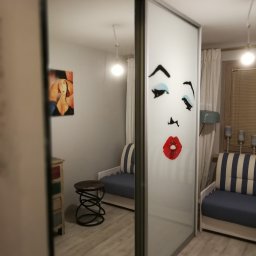 PPHU ROBMAX MEBLE - Szafa przesuwna z grafiką twarzy w pokoju z drewnianą podłogą. Widoczne meble, lampy i obraz na ścianie. Oświetlenie ciepłe, wnętrze przytulne.