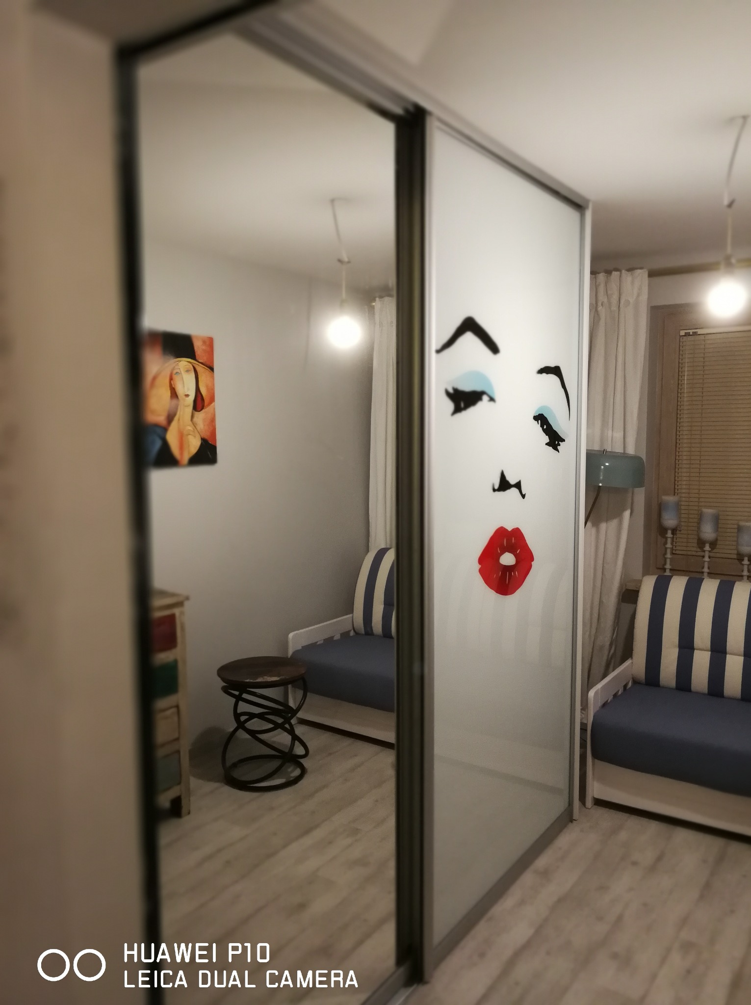 Szafa przesuwna z grafiką twarzy w pokoju z drewnianą podłogą. Widoczne meble, lampy i obraz na ścianie. Oświetlenie ciepłe, wnętrze przytulne.