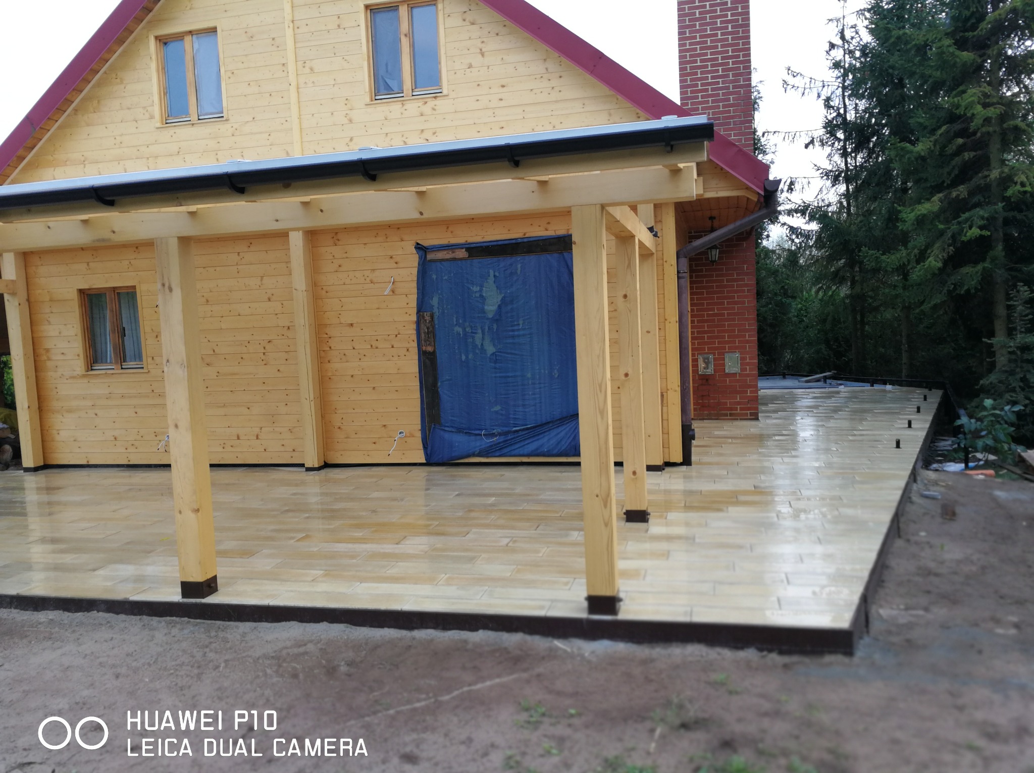 Wykończony taras z jasnych płytek przy drewnianym domu z elementami z cegły. Widoczna konstrukcja zadaszenia tarasu z drewnianych belek.