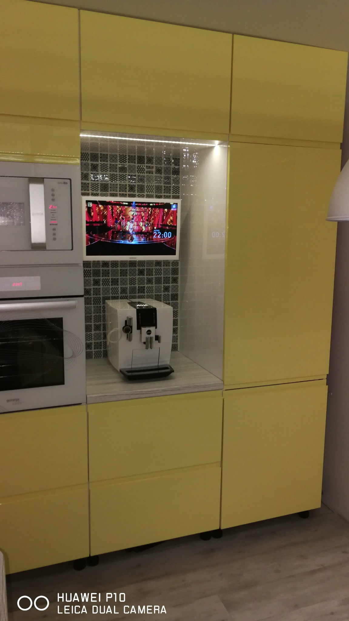 Żółta zabudowa kuchenna z piekarnikiem, mikrofalówką i ekspresem do kawy. Nad ekspresem podświetlona mozaika i telewizor. Nowoczesny design.