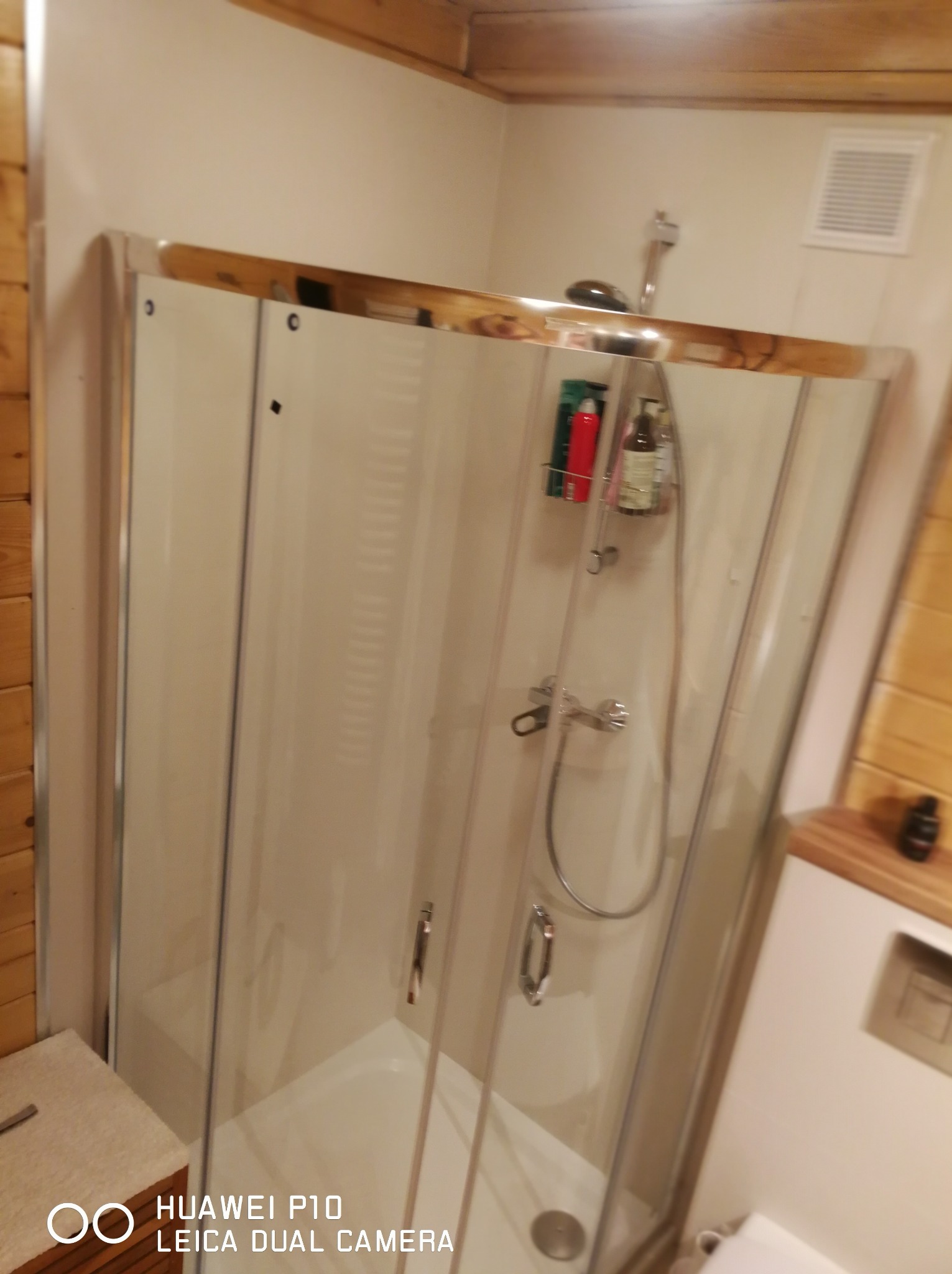 Kabina prysznicowa z przesuwnymi drzwiami w łazience wykończonej drewnem, widoczna armatura i półka z kosmetykami. Ujęcie z bliska, lekko zniekształcone perspektywą.