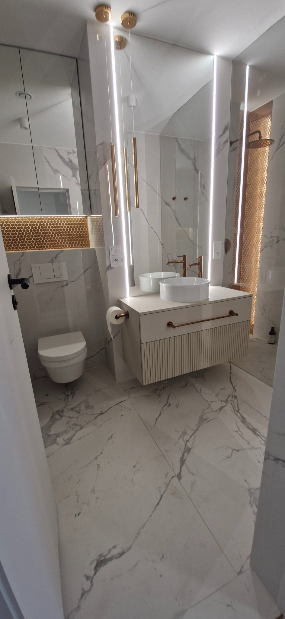 Nowoczesna łazienka z marmurową podłogą i ścianami. Umywalka nablatowa na szafce z lamelami, złote akcenty, oświetlenie LED i dekoracyjna mozaika w strefie prysznica.