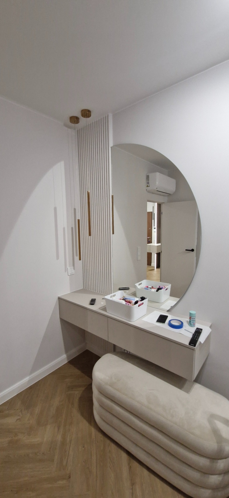 Nowoczesna toaletka z okrągłym lustrem i dekoracyjną ścianką z lamelami, oświetlenie punktowe, drewniana podłoga w jodełkę, kremowy puf. Minimalistyczny design.
