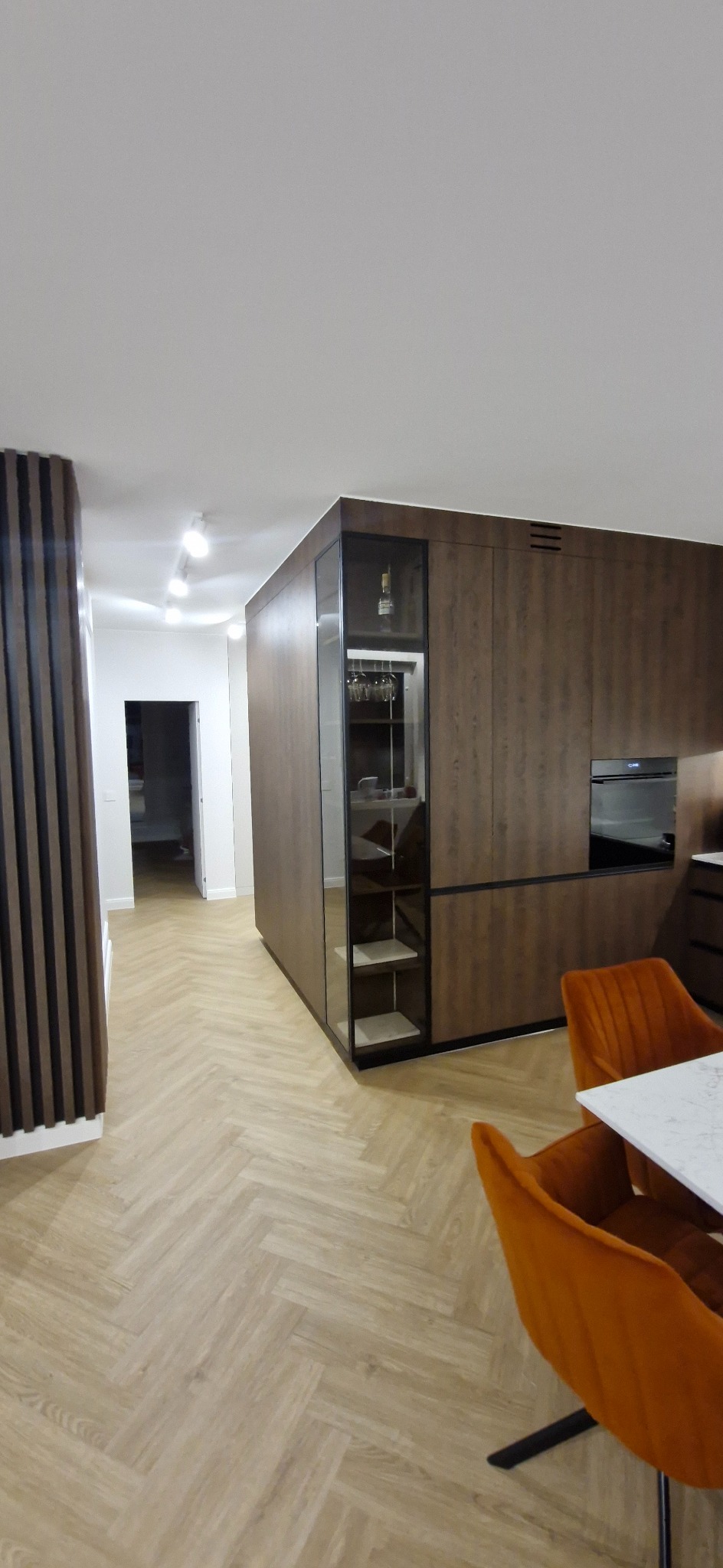 Nowoczesna kuchnia z zabudową w odcieniach brązu, parkietem w jodełkę i pomarańczowymi krzesłami. Widoczny fragment salonu z dekoracyjną ścianką z lameli.