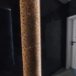 Nowoczesna ściana z podświetlanymi heksagonalnymi płytkami w odcieniach brązu, sąsiadująca z czarną, błyszczącą powierzchnią i fragmentem białych drzwi.