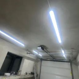 Wnętrze garażu z zamontowaną białą, segmentową bramą garażową, widoczne prowadnice i napęd. Oświetlenie stanowią długie lampy LED. W tle okno i drabina.