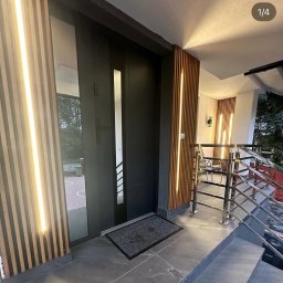 Elektryk Kacper Działo - Nowoczesne wejście do domu z czarnymi drzwiami, szarą podłogą i drewnianymi panelami z pionowym oświetleniem LED po bokach.