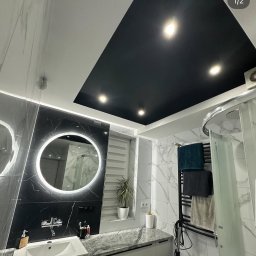 Elektryk Kacper Działo - Nowoczesna łazienka z czarnym sufitem podwieszanym i okrągłym lustrem LED. Marmurowe płytki, biała szafka pod umywalką i grzejnik drabinkowy.