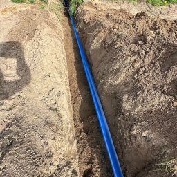 Elektryk Kacper Działo - Wkopana niebieska rura w ziemi, prawdopodobnie do instalacji elektrycznej na zewnątrz. Widoczny fragment pracownika w tle i trawa z boku wykopu.