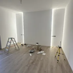 Prace elektryczne w nowym wnętrzu w Dąbrówce Wisłockiej: widoczne kable, puszki elektryczne, poziomica laserowa i drabina. Przygotowanie do montażu oświetlenia.