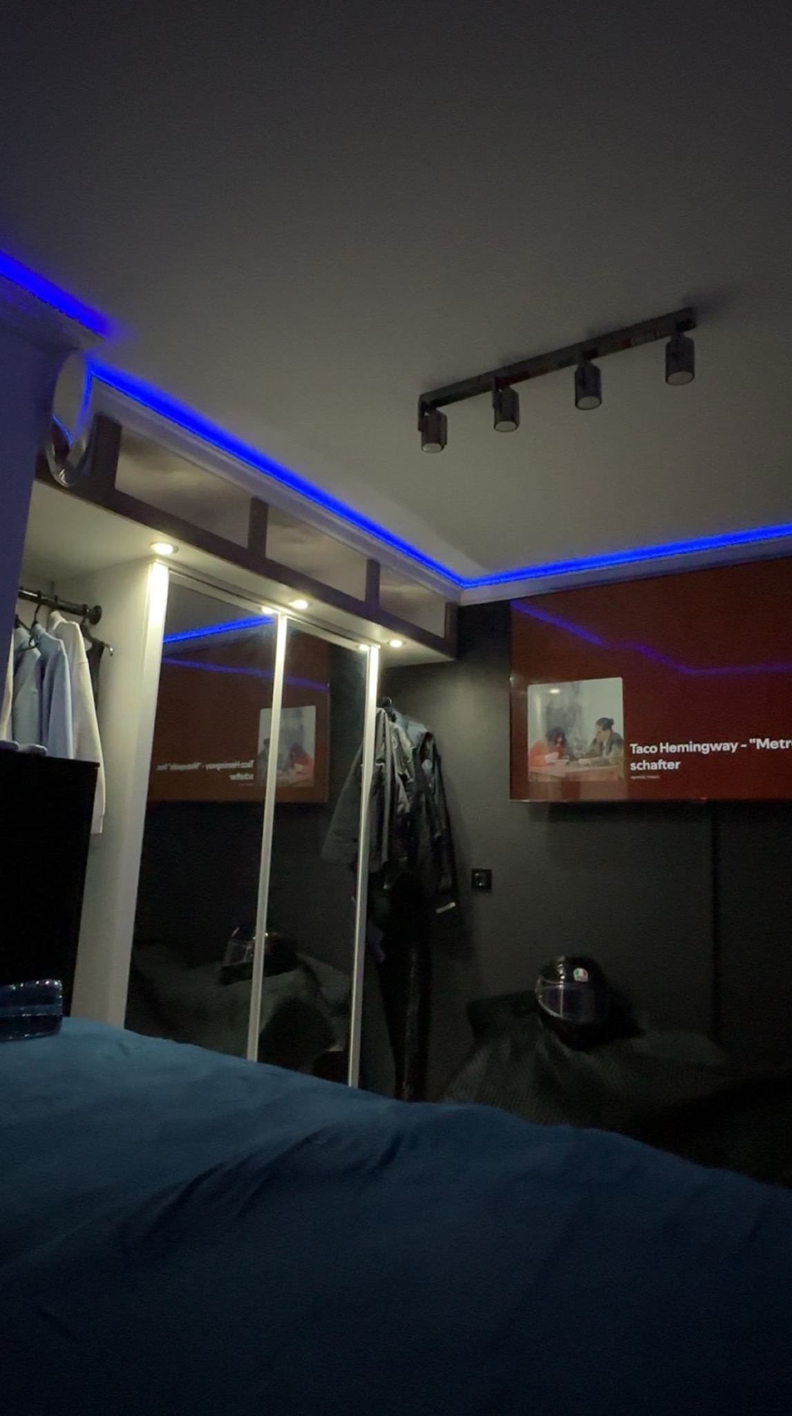 Nowoczesna sypialnia z czarnymi ścianami, podświetleniem LED i szafą z lustrzanymi drzwiami. Na ścianie wisi telewizor. Widoczne oświetlenie sufitowe.