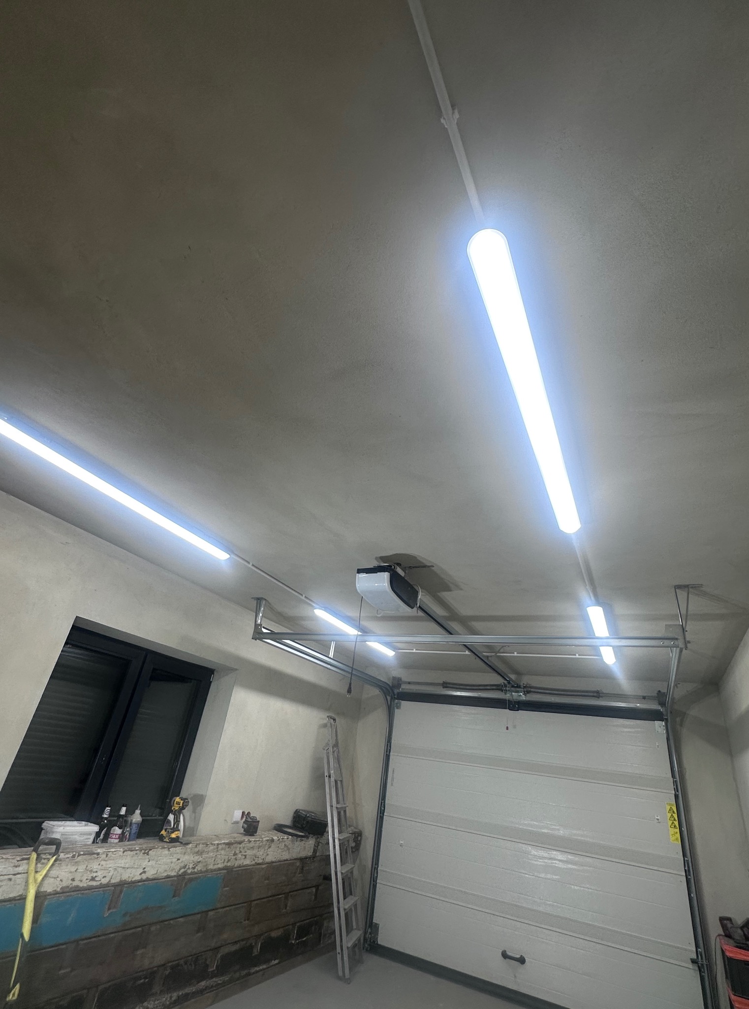 Wnętrze garażu z zamontowaną białą, segmentową bramą garażową, widoczne prowadnice i napęd. Oświetlenie stanowią długie lampy LED. W tle okno i drabina.