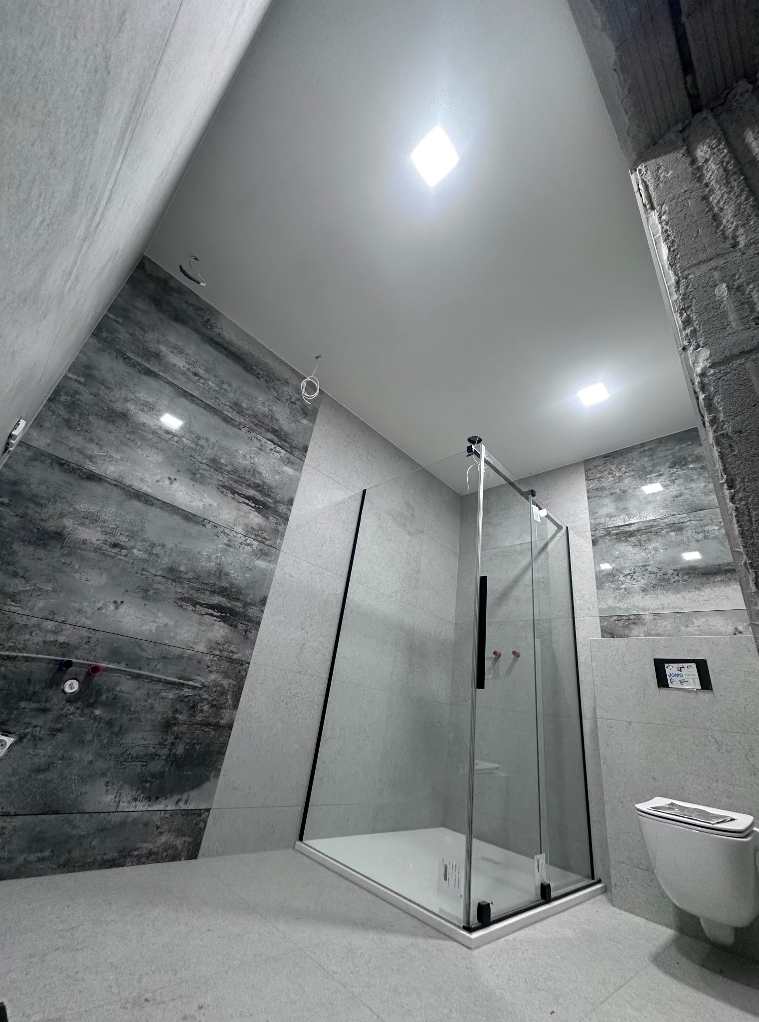 Nowoczesna kabina prysznicowa z grafitową ścianą w wykończonej łazience, widok z dołu, kwadratowe oświetlenie sufitowe, wisząca toaleta.
