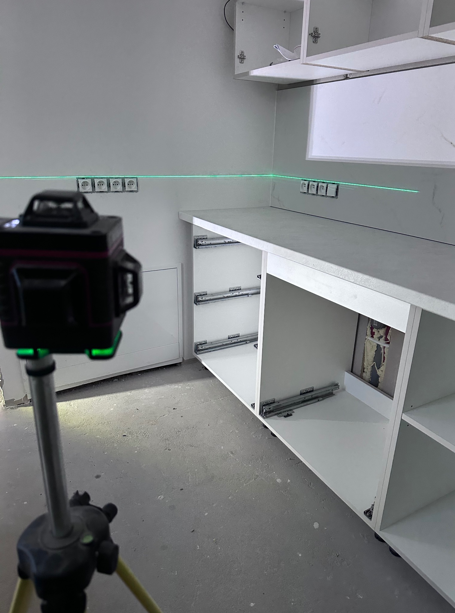 Poziomowanie laserowe gniazdek elektrycznych w nowej, białej zabudowie kuchennej. Widoczne szyny do szuflad. Precyzyjny montaż instalacji.
