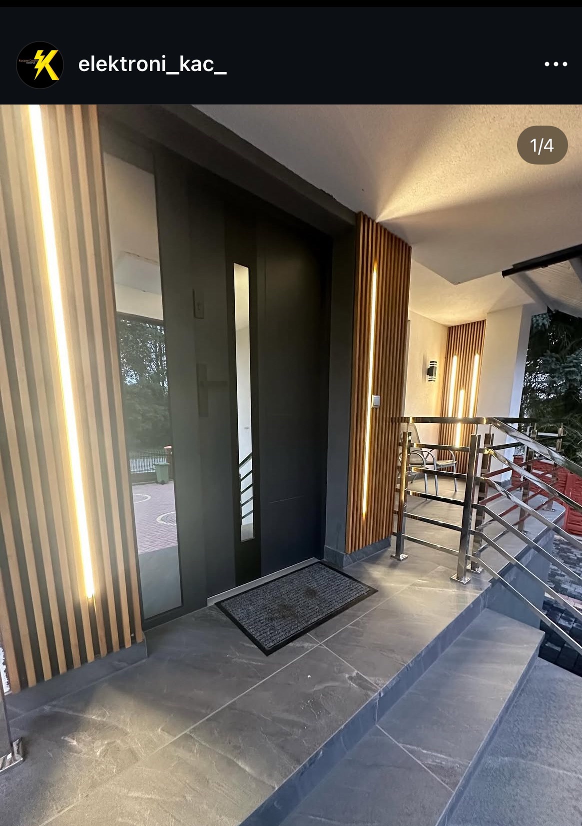 Nowoczesne wejście do domu z czarnymi drzwiami, szarą podłogą i drewnianymi panelami z pionowym oświetleniem LED po bokach.