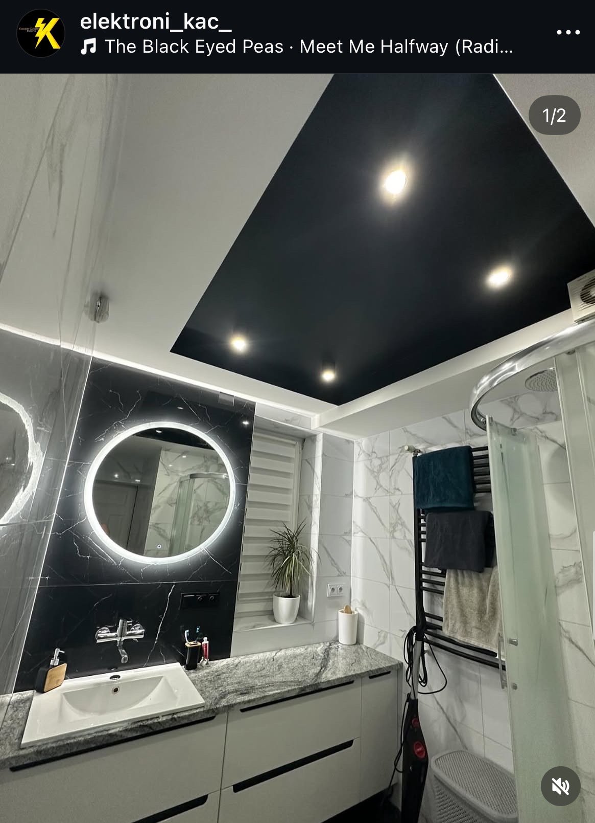 Nowoczesna łazienka z czarnym sufitem podwieszanym i okrągłym lustrem LED. Marmurowe płytki, biała szafka pod umywalką i grzejnik drabinkowy.