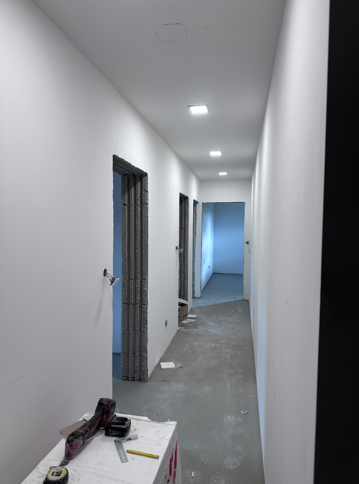 Wnętrze korytarza w trakcie wykańczania: białe ściany, surowy beton wokół otworów drzwiowych, widoczne narzędzia na pierwszym planie, sufit z oświetleniem.