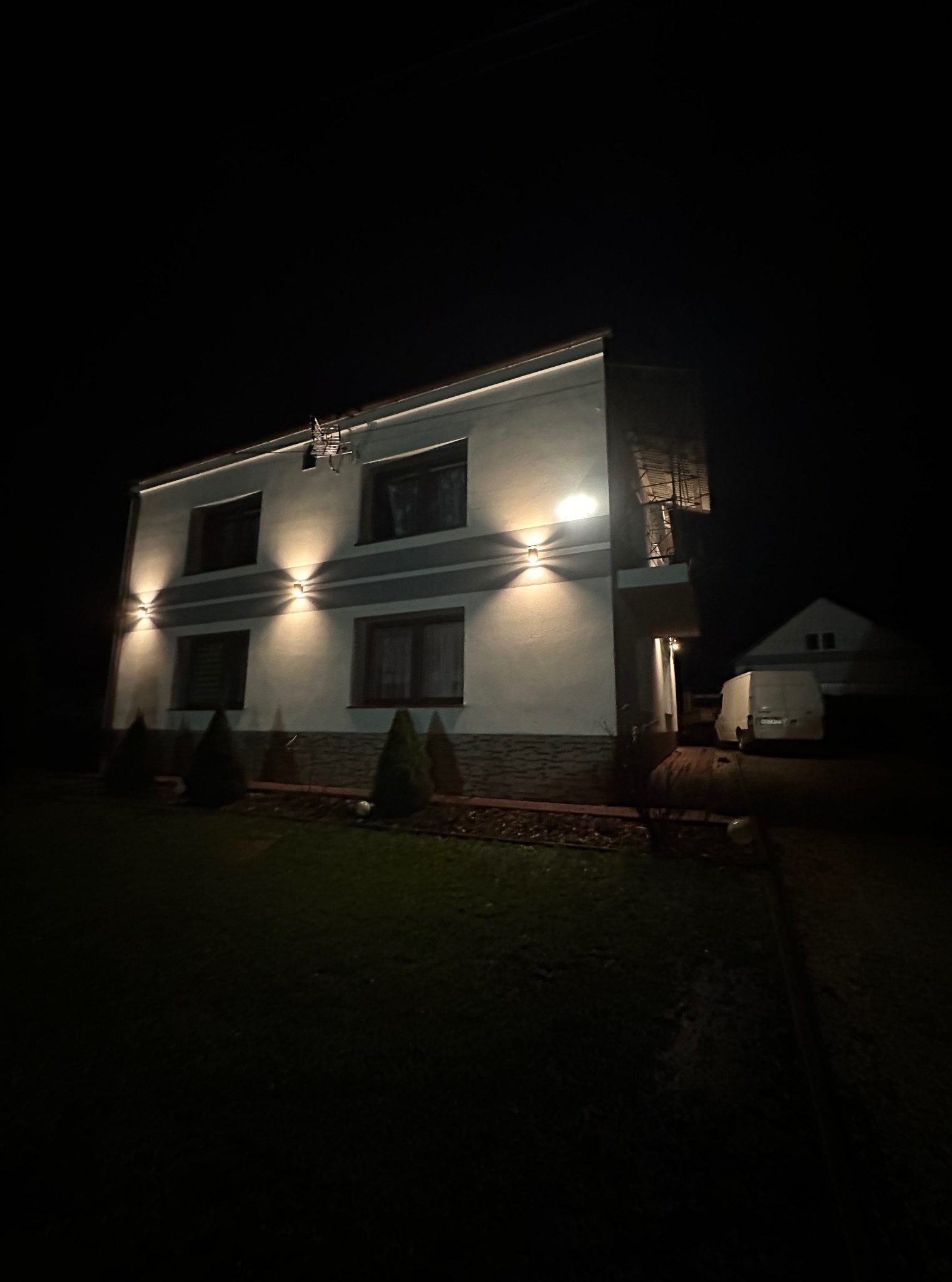 Dwupiętrowy dom z oświetleniem elewacyjnym w nocy. Widoczne lampy ścienne i listwa LED pod dachem. Na posesji zaparkowany biały van.