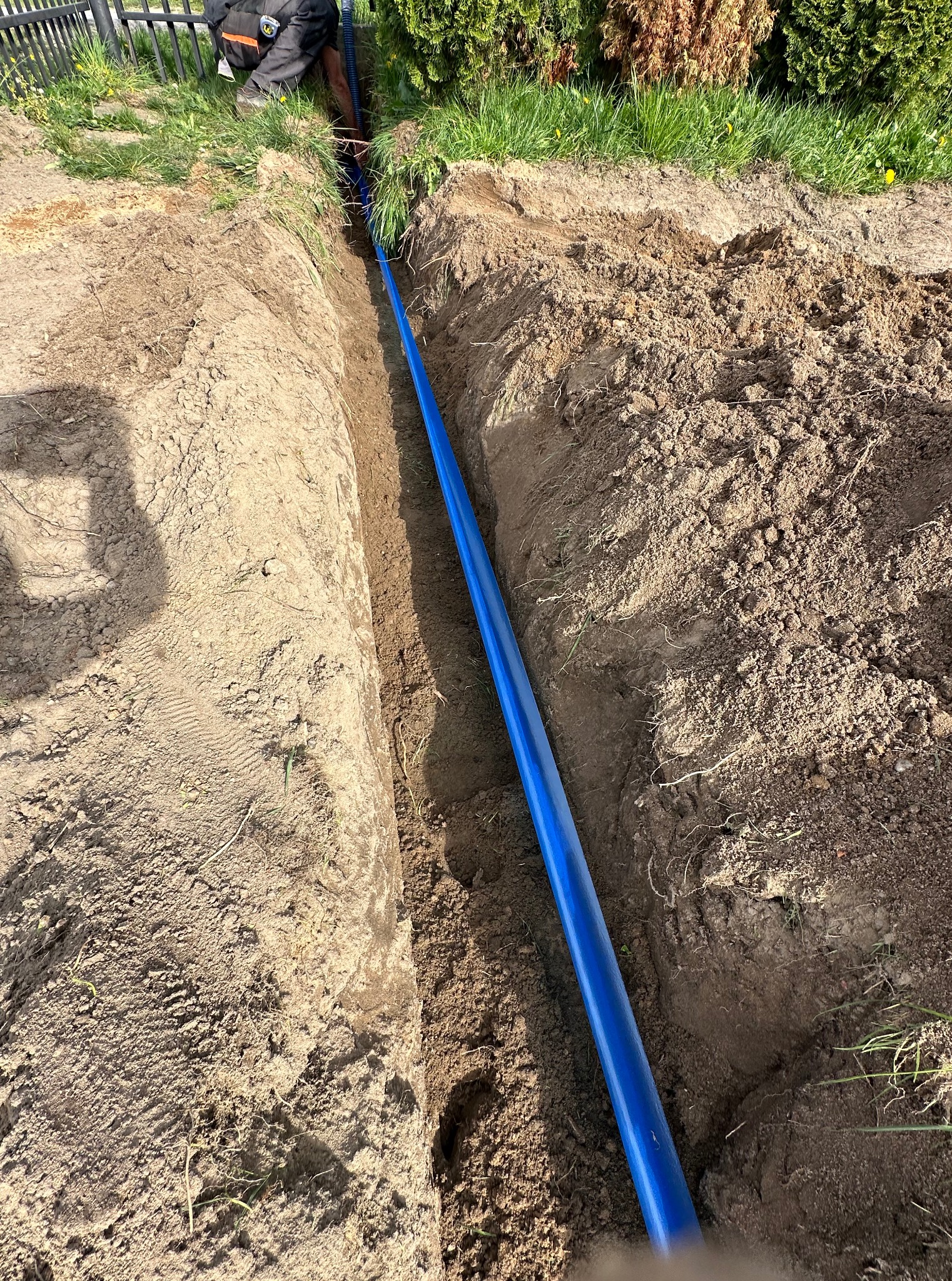 Wkopana niebieska rura w ziemi, prawdopodobnie do instalacji elektrycznej na zewnątrz. Widoczny fragment pracownika w tle i trawa z boku wykopu.