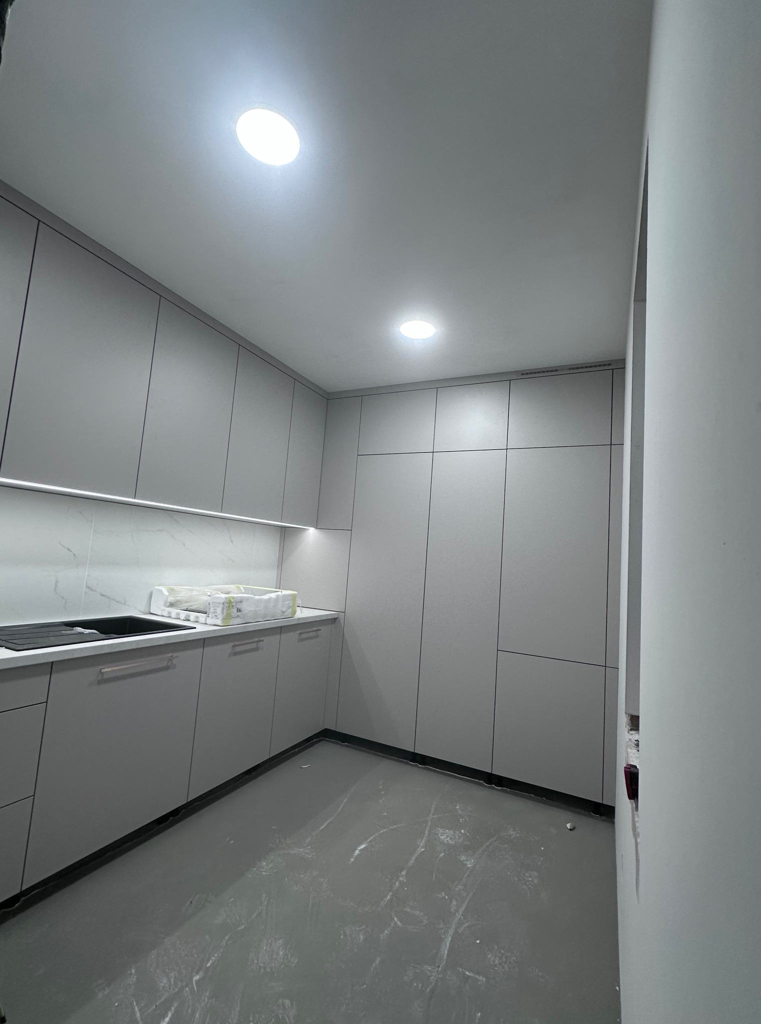 Nowoczesna, szara kuchnia z oświetleniem LED pod szafkami, marmurowym backsplashiem i błyszczącą podłogą. Minimalistyczny design z widocznymi elementami wykończeniowymi.