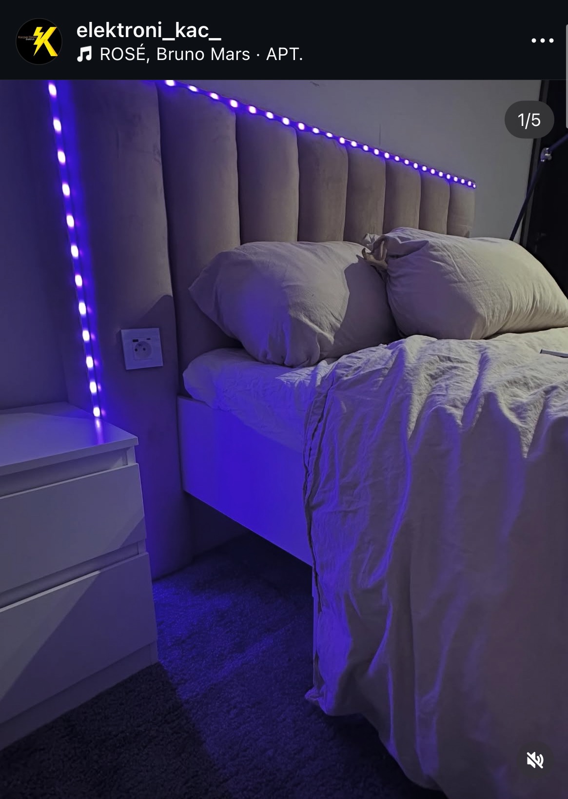 Podświetlane łóżko z wezgłowiem w sypialni. Taśma LED tworzy nastrojowe oświetlenie. Widoczny fragment szafki nocnej i gniazdko elektryczne na ścianie.