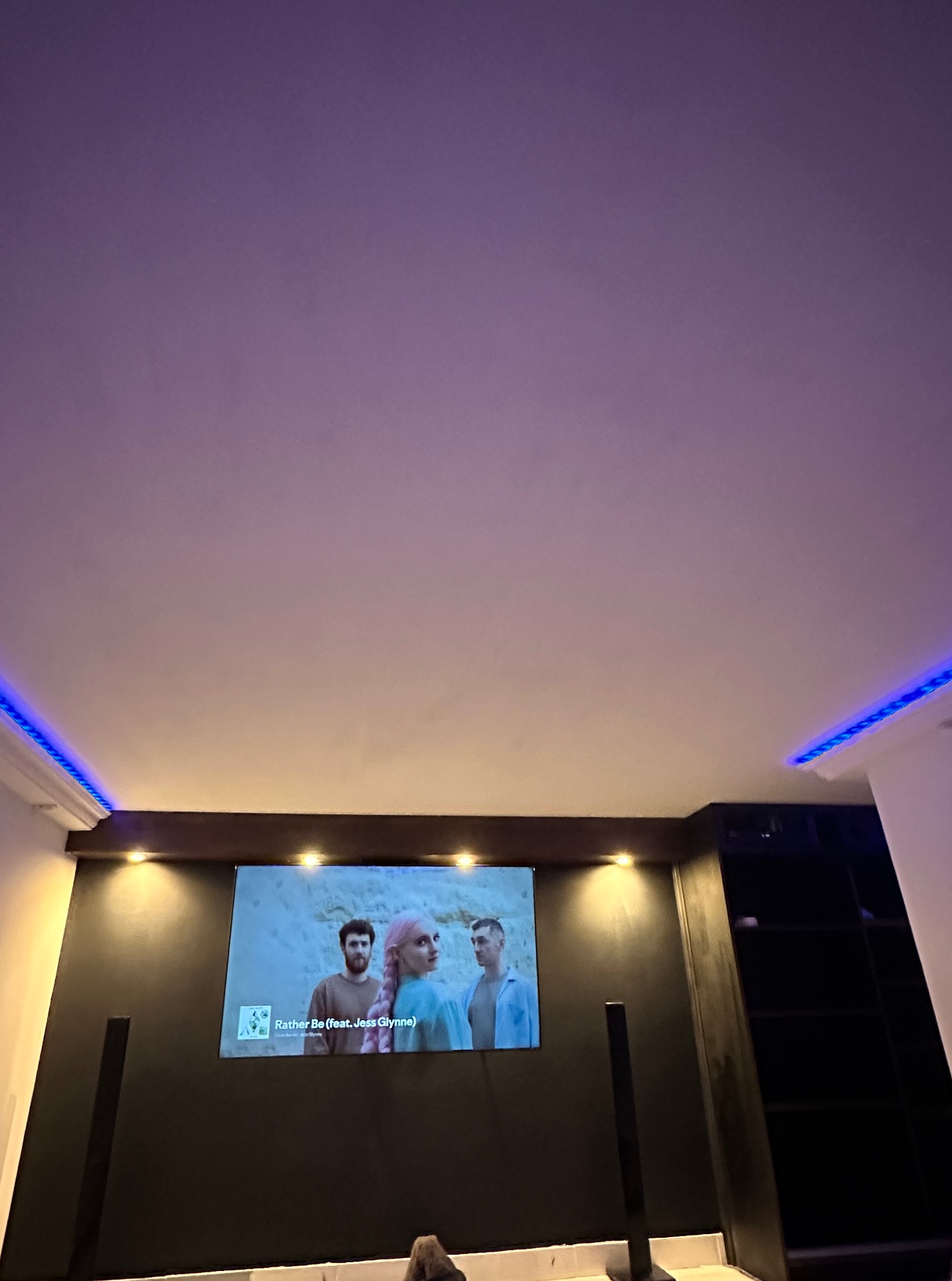 Nowoczesny salon z podświetlanym sufitem LED i wbudowanym telewizorem, tworzący przytulną atmosferę. Minimalistyczny design z ciemną ścianą i jasnym sufitem.