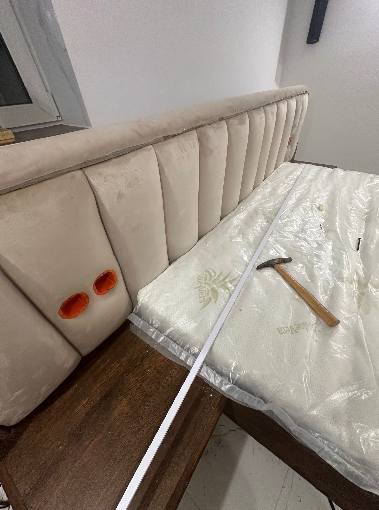 Montaż łóżka z tapicerowanym zagłówkiem, widoczne narzędzia i listwa montażowa na materacu w folii. Dwa pomarańczowe elementy na zagłówku.