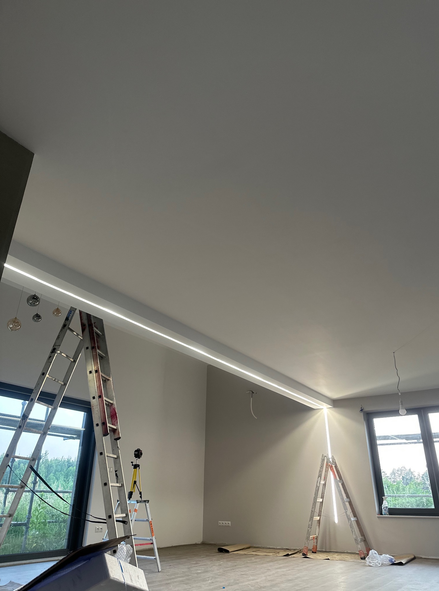 Nowoczesne oświetlenie LED w suficie podwieszanym, widoczne drabiny monterskie i laser krzyżowy, szare ściany, okna z widokiem na zieleń. Montaż oświetlenia.