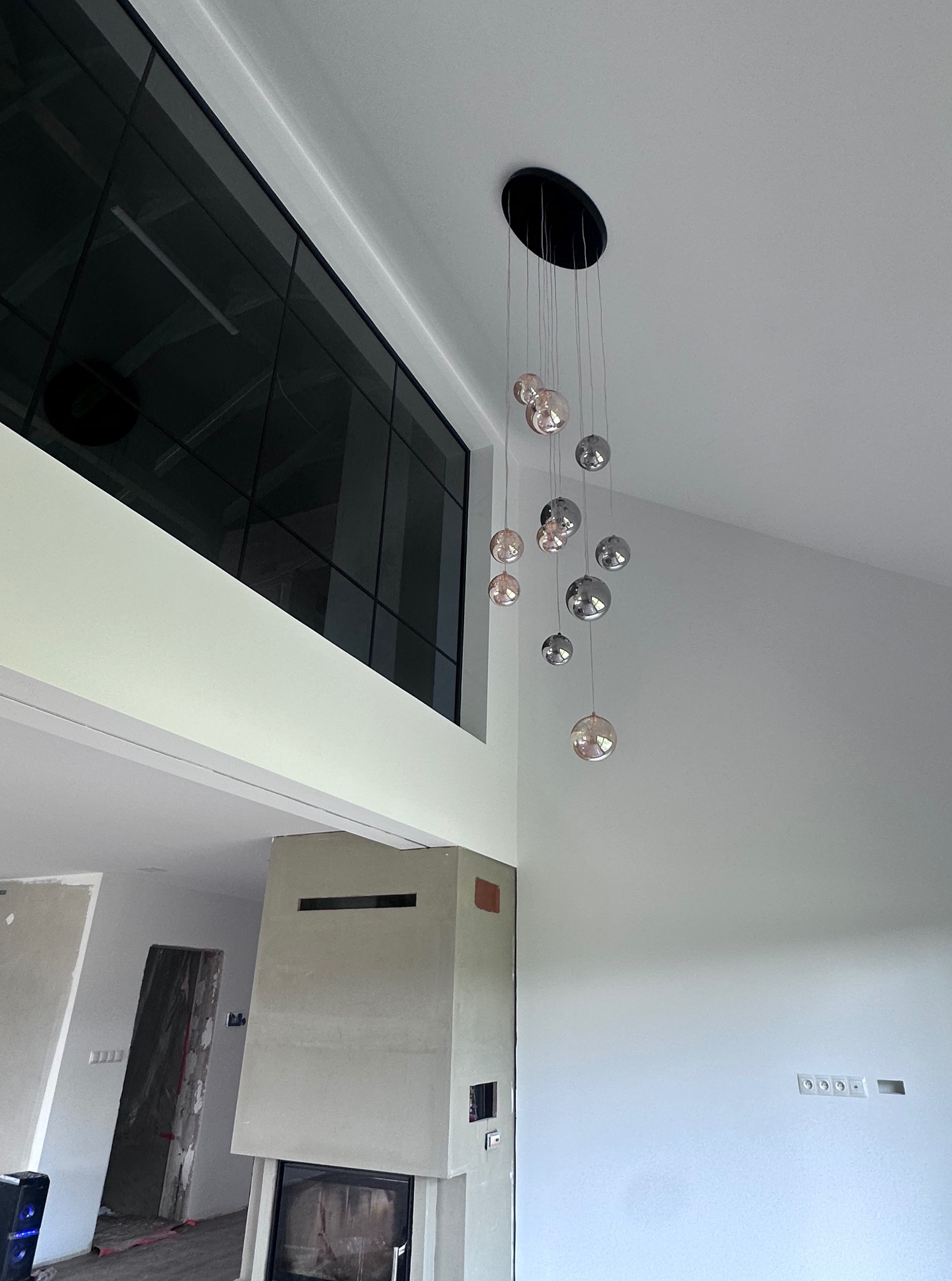 Nowoczesny żyrandol kulisty w przestronnym wnętrzu z dużym oknem i kominkiem. Minimalistyczny design, surowe ściany, widoczne gniazdka elektryczne.