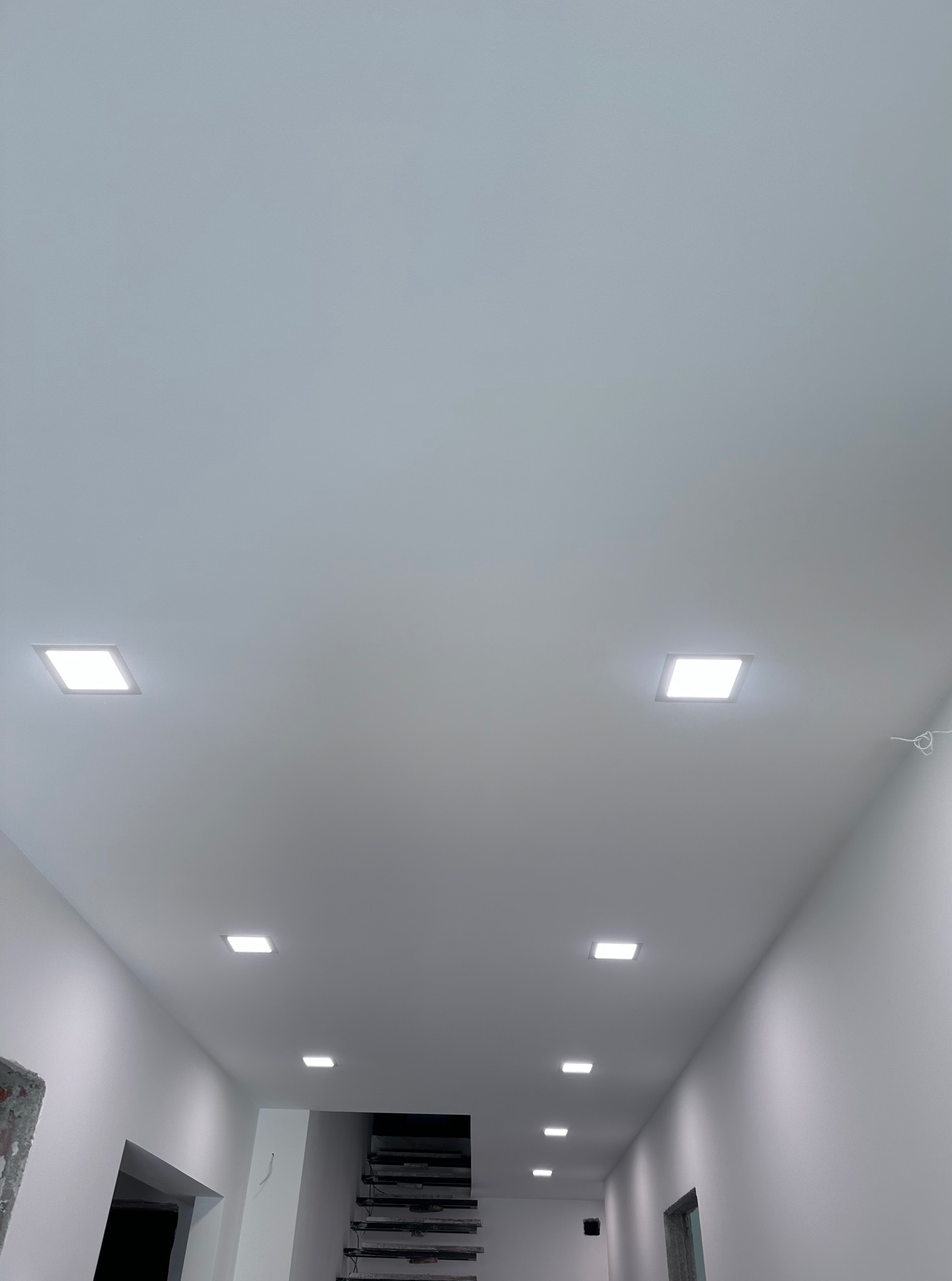 Biały sufit z równomiernie rozmieszczonymi kwadratowymi lampami LED, oświetlającymi korytarz w nowym budynku. Widoczne schody w głębi.