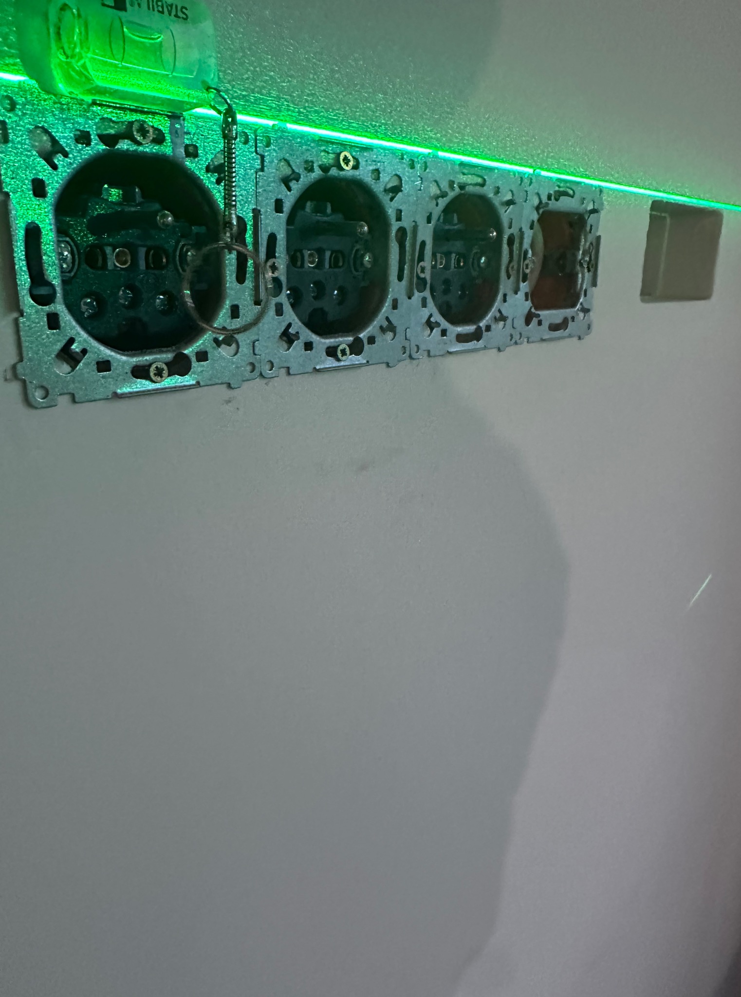 Potrójne gniazdko elektryczne podłączone do ściany, poziomowane laserowo. Widoczne metalowe elementy montażowe i zielona linia lasera.