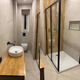 EMI Butik Kwiatowy Ewa Misiak - Nowoczesna łazienka po remoncie: biała umywalka na drewnianym blacie, kabina prysznicowa z czarną ramą, beżowe płytki i drewniane akcenty na ścianach.