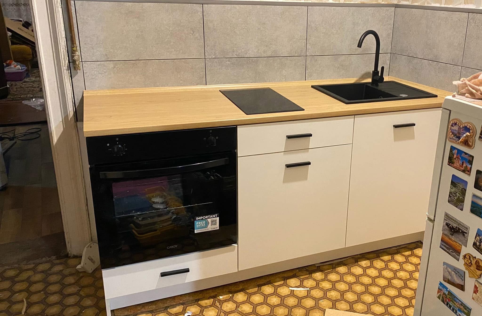 Nowoczesna, jasna kuchnia z drewnianym blatem, czarnym zlewem i płytą grzewczą. Uwagę przyciąga kontrast między nowym wyposażeniem a wzorzystą podłogą.