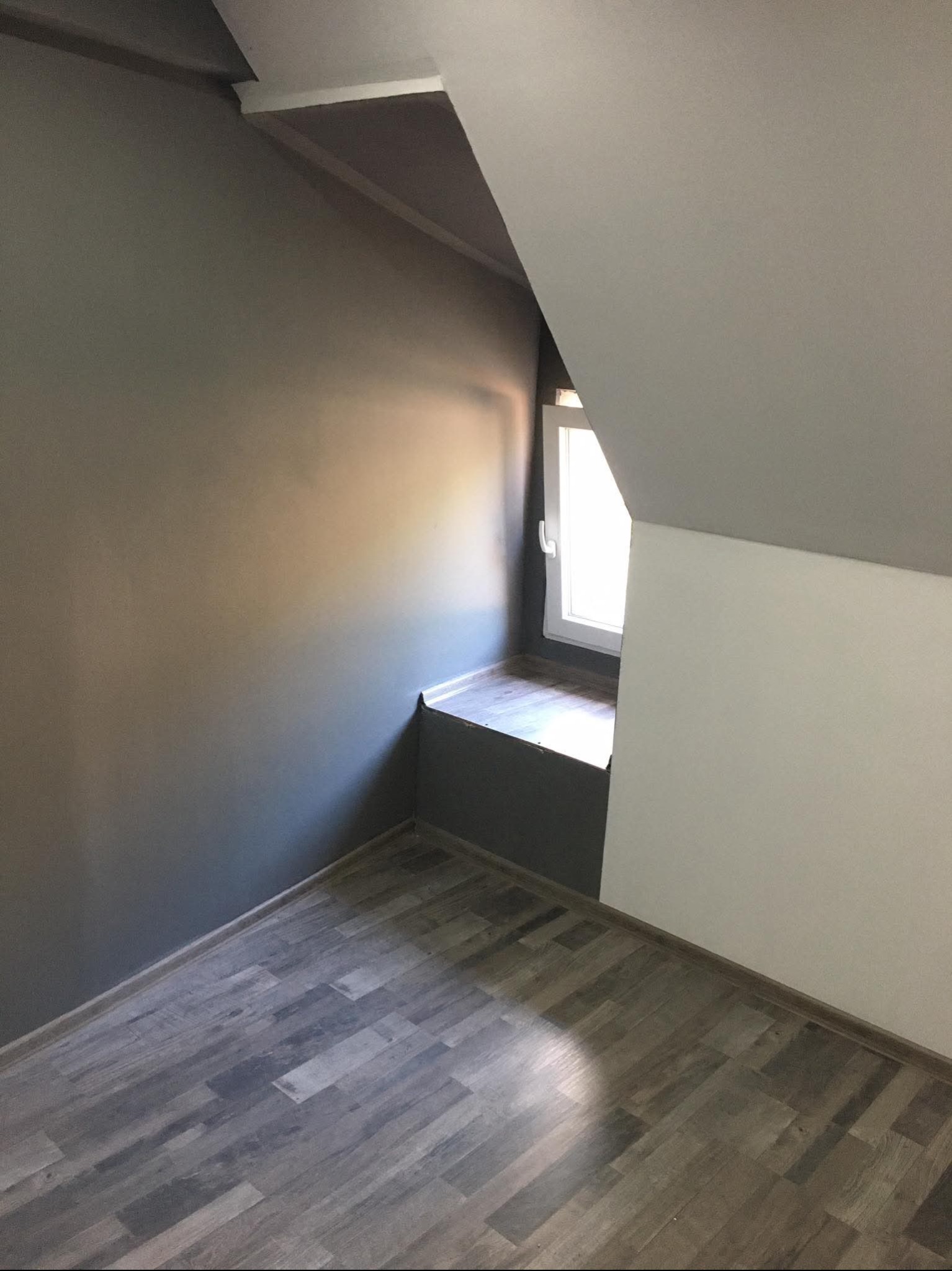 Pokój na poddaszu po malowaniu ścian na szaro i biało, z drewnianą podłogą i oknem wnękowym. Świeżo wykończone wnętrze z widoczną strukturą drewna.