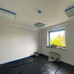 Kacper Malinowski - Białe, odmalowane wnętrze pokoju zabezpieczone niebieską taśmą malarską i folią ochronną na podłodze. Widoczne okno z zielenią za szybą i narzędzia malarskie.