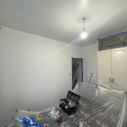 Kacper Malinowski - Pokój w trakcie malowania w Słupsku. Zabezpieczone meble folią, ściany świeżo pomalowane na biało, widoczna skrzynka z narzędziami.