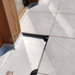 Kacper Malinowski - Montaż płytek na balkonie z drewnianą ramą okienną. Widoczne podkładki pod płytkami. Jasne, równe płytki tworzą nowoczesną powierzchnię.