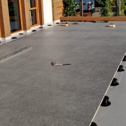 Kacper Malinowski - Układanie szarych płytek na balkonie z użyciem regulowanych wsporników. Widoczny poziomica i narzędzia. Częściowo ukończona podłoga na zewnątrz budynku.