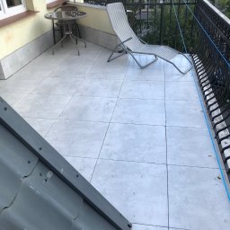 Kacper Malinowski - Jasny balkon z szarymi płytkami, metalowym stolikiem i leżakiem. Balustrada z ozdobnymi elementami. Fragment dachu w rogu kadru. Widok na zieleń i zabudowę.