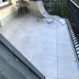 Jasny balkon z szarymi płytkami, metalowym stolikiem i leżakiem. Balustrada z ozdobnymi elementami. Fragment dachu w rogu kadru. Widok na zieleń i zabudowę.