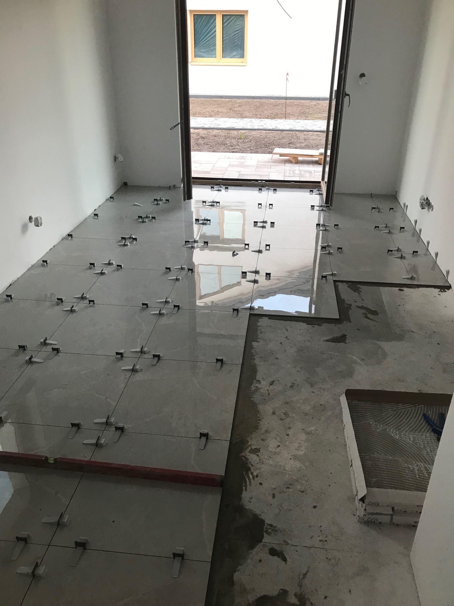 Układanie płytek podłogowych w jasnym pomieszczeniu z otwartym wyjściem na zewnątrz, poziomica i krzyżyki dystansowe widoczne na powierzchni płytek.