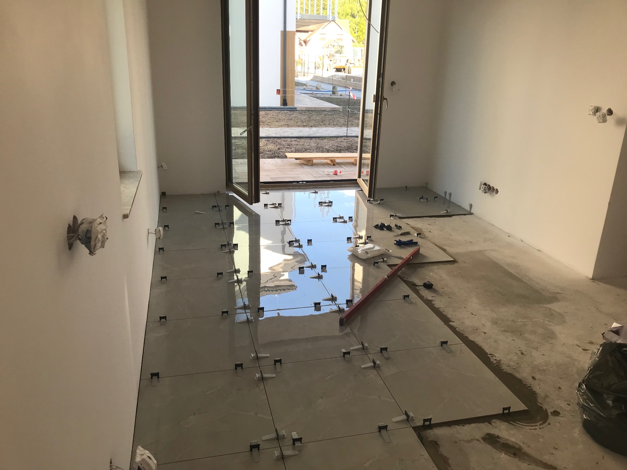 Jasne płytki podłogowe w trakcie układania, z użyciem klipsów poziomujących, widok na otwarte drzwi balkonowe i zewnętrzny taras w tle. Częściowo wykończone ściany.