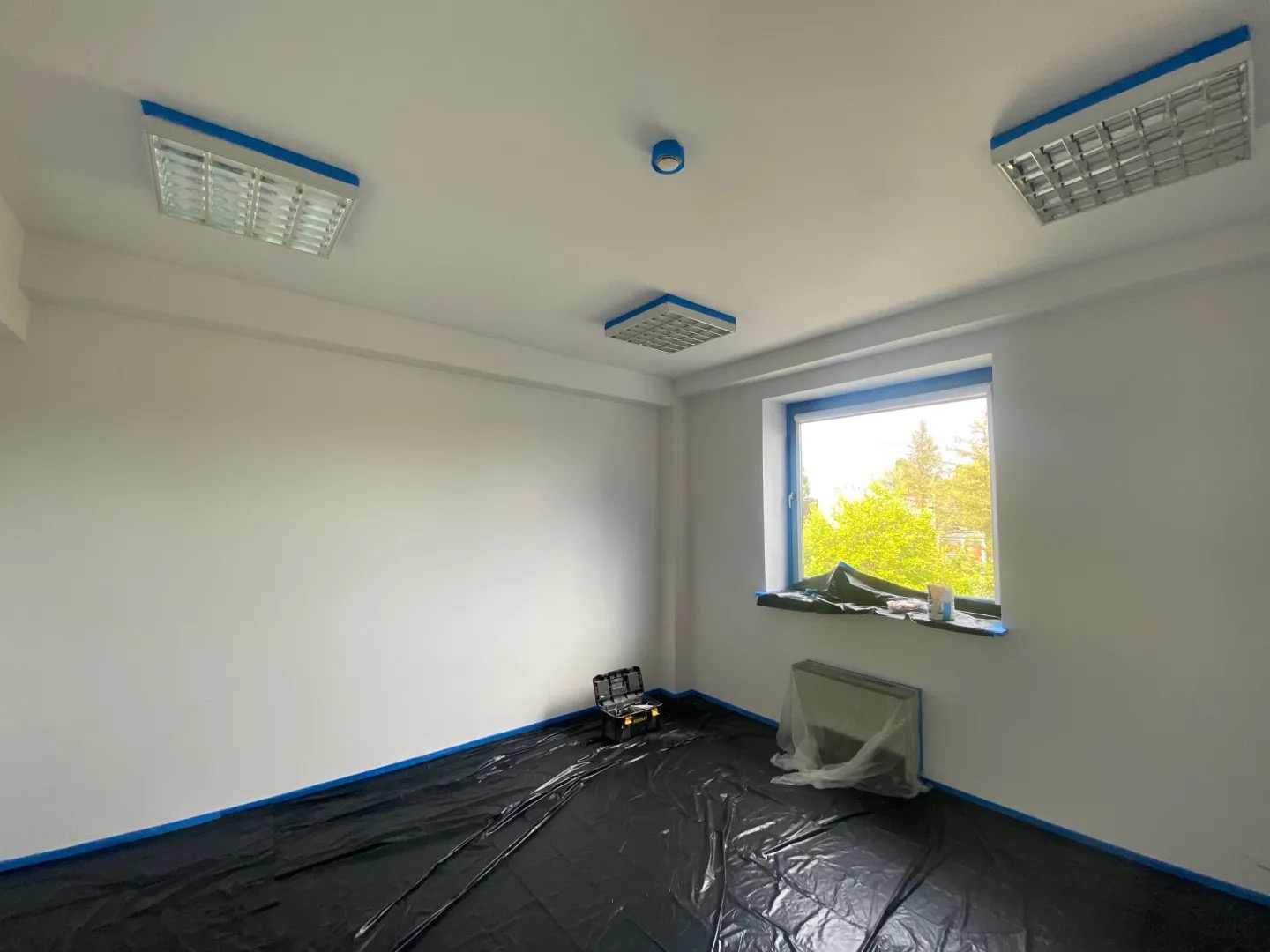 Białe, odmalowane wnętrze pokoju zabezpieczone niebieską taśmą malarską i folią ochronną na podłodze. Widoczne okno z zielenią za szybą i narzędzia malarskie.