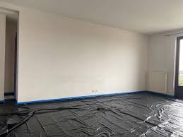 Pokój przygotowany do malowania: folia ochronna na podłodze, taśma malarska przy ścianach. Widok na jasne ściany i okno balkonowe.