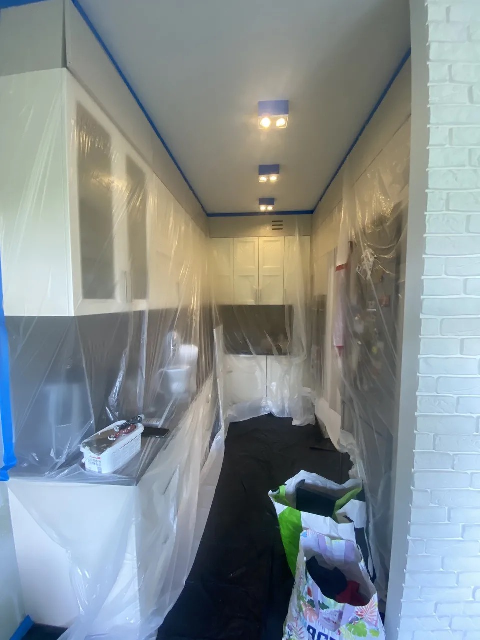 Kuchnia zabezpieczona folią i taśmą malarską przed remontem. Widoczne białe szafki i czarne blaty. Podłoga zakryta czarną folią. Przygotowania do malowania.