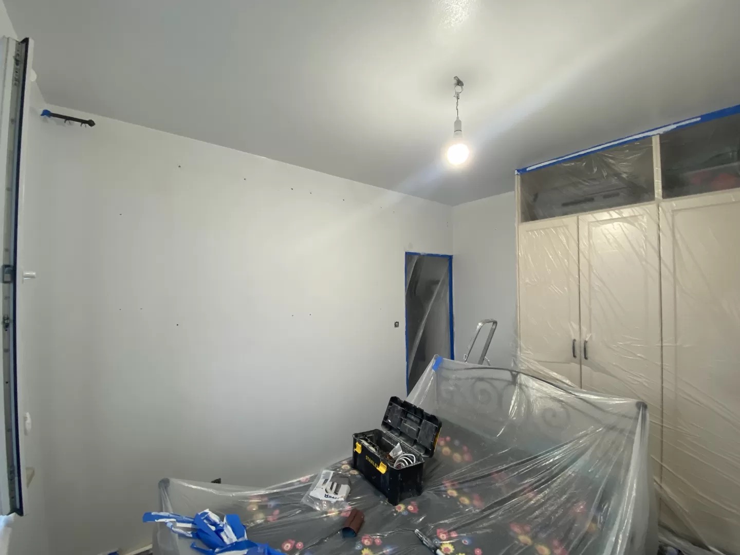 Pokój w trakcie malowania w Słupsku. Zabezpieczone meble folią, ściany świeżo pomalowane na biało, widoczna skrzynka z narzędziami.