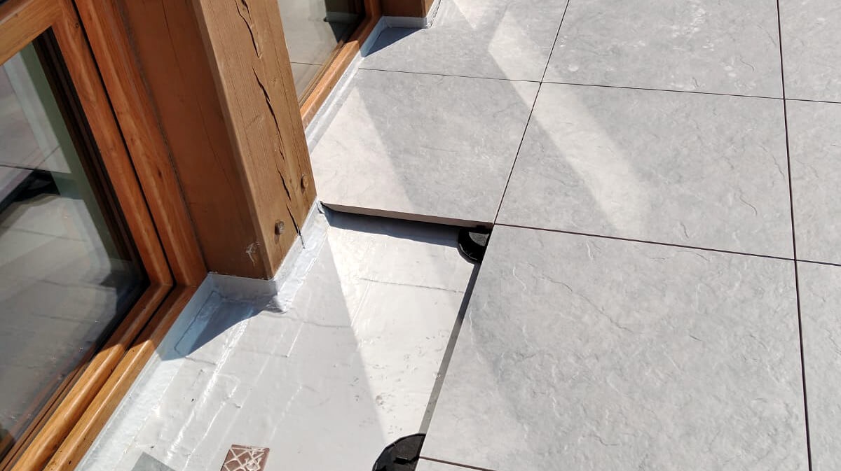 Montaż płytek na balkonie z drewnianą ramą okienną. Widoczne podkładki pod płytkami. Jasne, równe płytki tworzą nowoczesną powierzchnię.