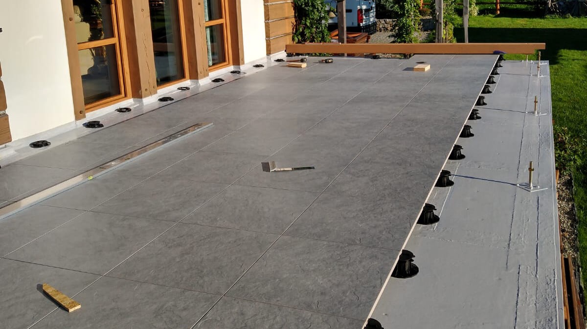 Układanie szarych płytek na balkonie z użyciem regulowanych wsporników. Widoczny poziomica i narzędzia. Częściowo ukończona podłoga na zewnątrz budynku.