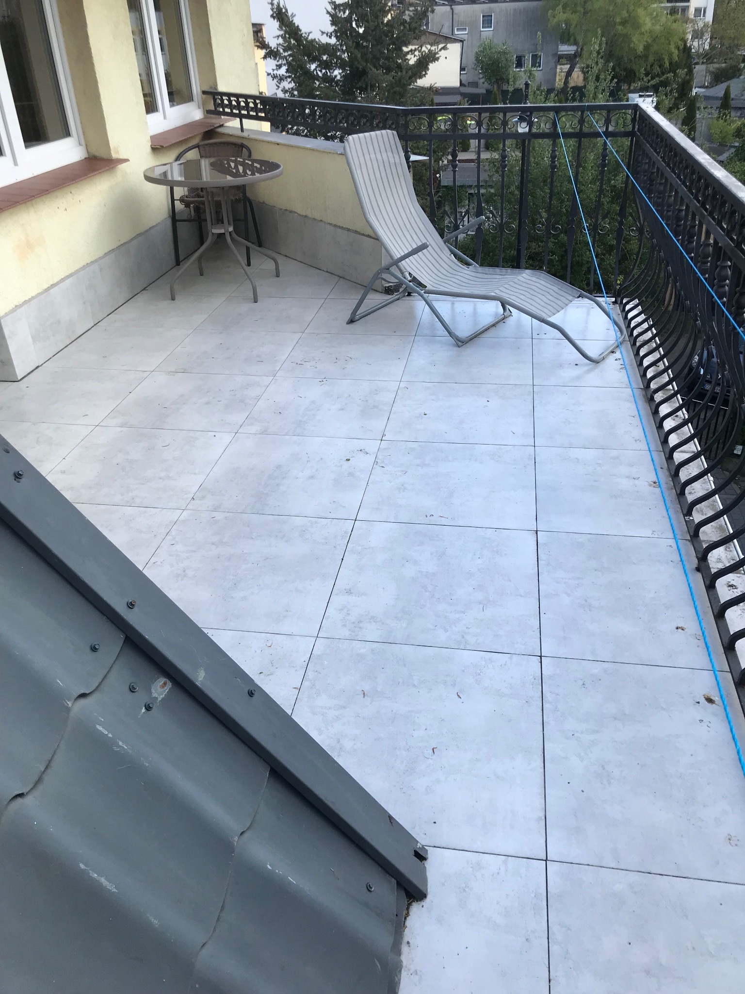 Jasny balkon z szarymi płytkami, metalowym stolikiem i leżakiem. Balustrada z ozdobnymi elementami. Fragment dachu w rogu kadru. Widok na zieleń i zabudowę.
